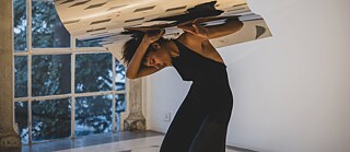 A dancing woman in a black dress holds a wavy mirror over her body with both hands. 